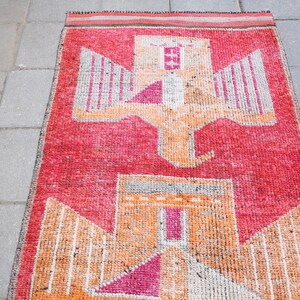 May include: A vintage rug with a geometric design featuring a stylized bird with outstretched wings. The rug is primarily red with orange and white accents.