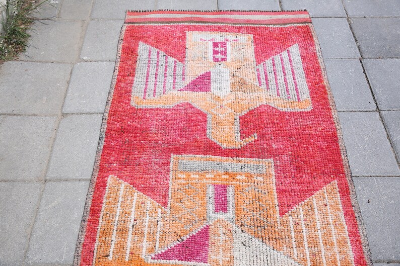 May include: A vintage rug with a geometric design featuring a stylized bird with outstretched wings. The rug is primarily red with orange and white accents.