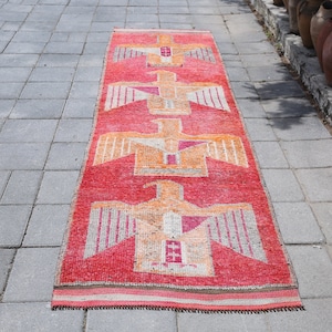 May include: A long, red runner rug with a repeating pattern of stylized birds in orange and white. The rug is laid out on a gray concrete patio.