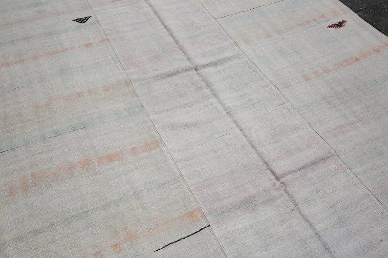 K&ouml;nnte beinhalten: Ein hellfarbiger, rechteckiger Teppich mit strukturierter Oberfl&auml;che. Der Teppich weist verblasste orangefarbene und schwarze horizontale Streifen und kleine geometrische Muster auf. Der Teppich besteht aus gewebtem Material.