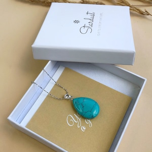 May include: A silver necklace with a large, teardrop-shaped turquoise pendant rests in an open white box. The box is on a tan surface, with a white lid nearby. The box is labeled "Stardust GIFTS FROM NATURE".