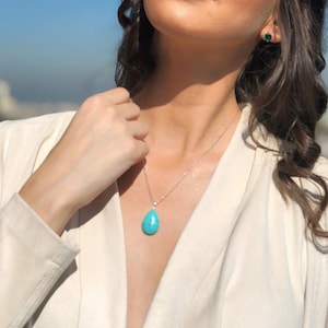 May include: A silver necklace with a turquoise teardrop pendant and a small diamond accent. The image also shows a pair of green gemstone stud earrings. The person is wearing a cream-colored blazer.