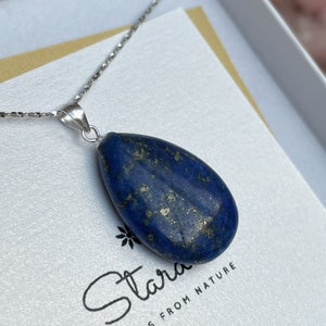 Può includere: Una collana a catena d'argento con un ciondolo a forma di lacrima in lapislazzuli. Il ciondolo è di colore blu scuro con macchie dorate. La collana è esposta su uno sfondo bianco con il testo "Stara from Nature".