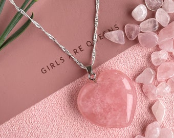 May include: A silver chain necklace with a large pink heart-shaped pendant made of rose quartz. The necklace is lying on a pink surface with a pink notebook that says "Girls are Queens" and several small rose quartz stones.