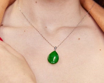 May include: A silver chain necklace with a green jade pendant in the shape of a teardrop. The necklace is worn by a person with a tan jacket and a white top. The person is also wearing a green jade bracelet.