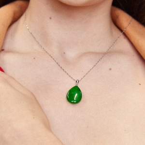 Peut inclure: Un collier en argent avec un pendentif en jade vert en forme de larme. Le collier est porté par une personne avec une veste beige et un haut blanc. La personne porte également un bracelet en jade vert.