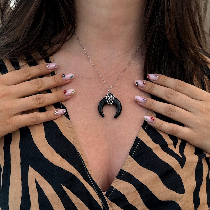 May include: A silver-toned chain necklace featuring a black crescent moon pendant. The pendant is accented with a small, triangular silver-toned cap embedded with tiny clear crystals at the top center, connecting the crescent to the chain.