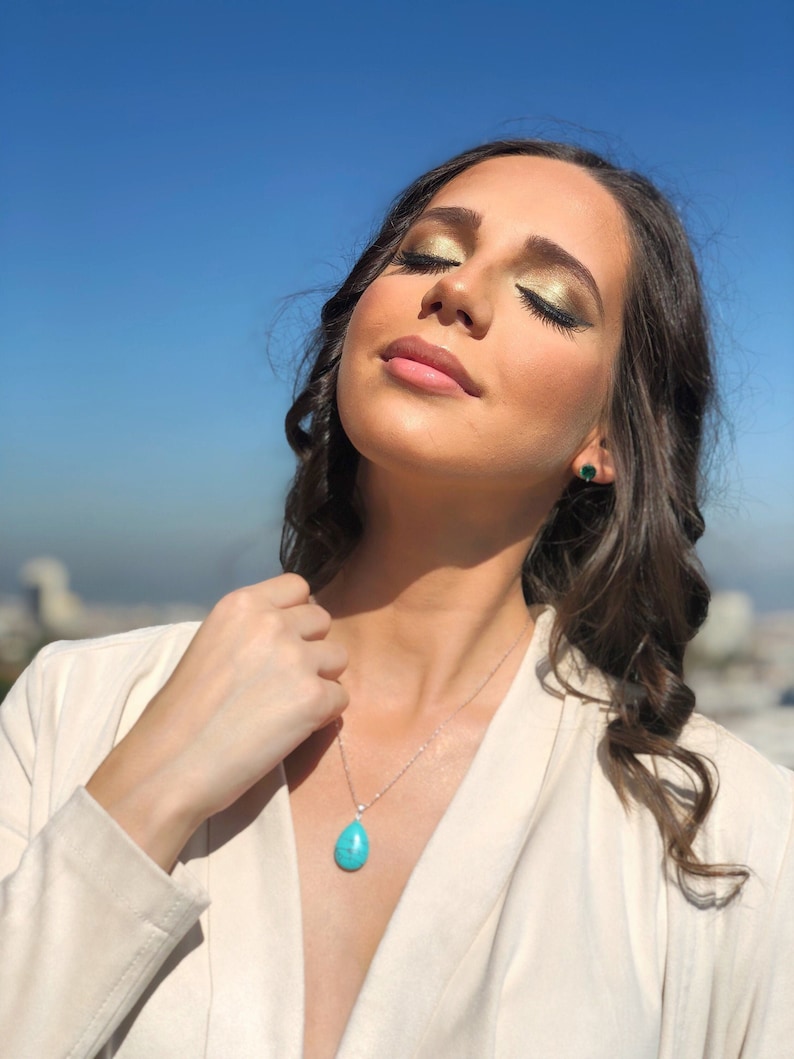 May include: A silver necklace with a turquoise teardrop pendant and a small diamond accent. The image also shows a pair of green gemstone stud earrings. The person is wearing a cream-colored blazer.