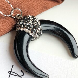 May include: A necklace featuring a black, crescent-shaped horn pendant topped with a cap of sparkling, dark gray rhinestones. The pendant hangs from a delicate silver-toned chain, creating a contrast between the smooth, glossy black surface and the faceted, reflective stones at the top.