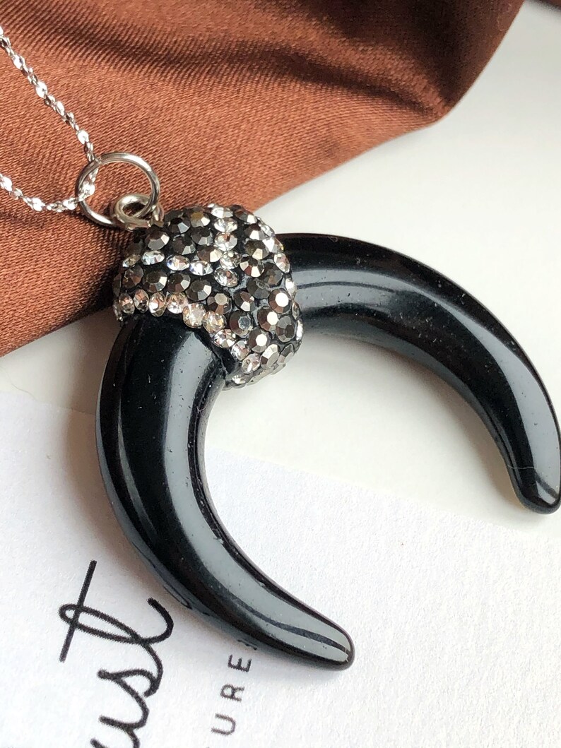 May include: A necklace featuring a black, crescent-shaped horn pendant topped with a cap of sparkling, dark gray rhinestones. The pendant hangs from a delicate silver-toned chain, creating a contrast between the smooth, glossy black surface and the faceted, reflective stones at the top.
