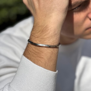 May include: A silver bangle bracelet with a flat, slightly curved design. The bracelet is worn on a person's wrist.