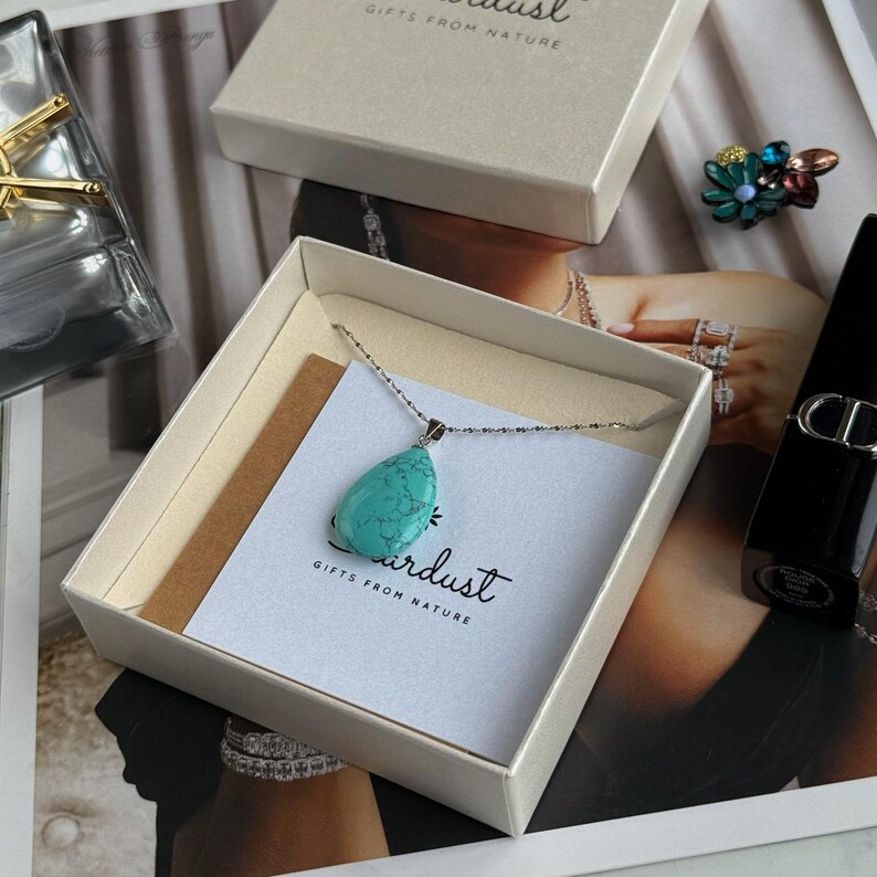 May include: A turquoise teardrop pendant necklace displayed in a gift box. The necklace has a silver chain and a large, polished turquoise stone. The box is beige, and a card with the text "Airdust Gifts From Nature" is visible.