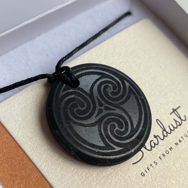 Puede incluir: Collar con colgante negro circular con un dise&ntilde;o de triple espiral. El colgante est&aacute; hecho de piedra negra pulida y cuelga de un cord&oacute;n negro. El texto "Stardust Gifts from Nature" es visible en el fondo.