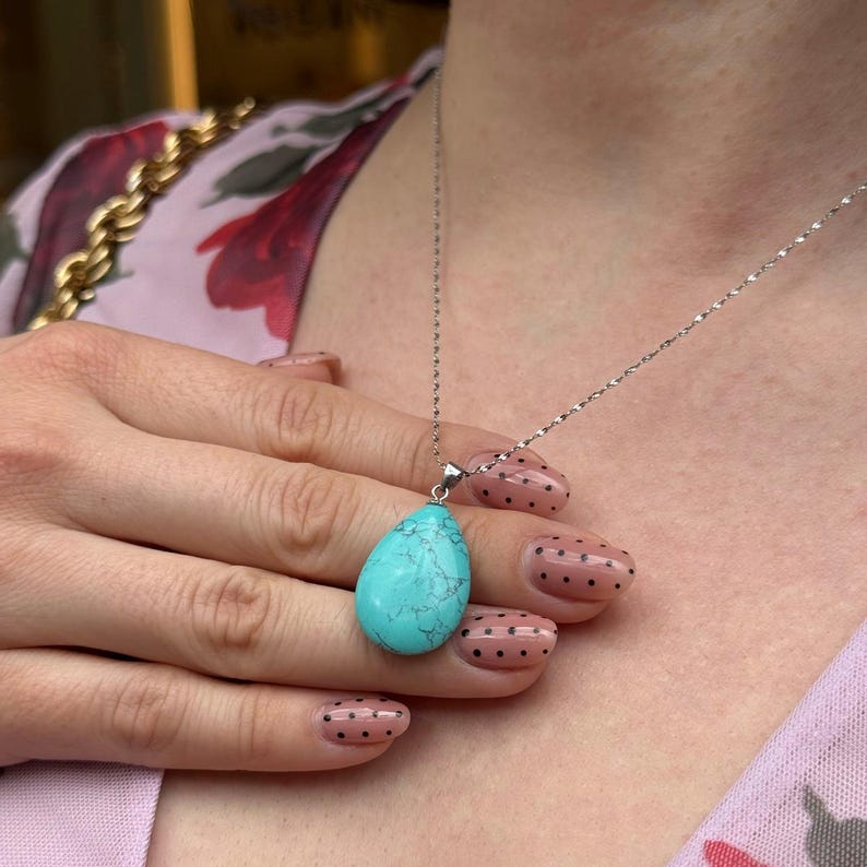 May include: A silver necklace featuring a teardrop-shaped turquoise pendant. The pendant has a marbled appearance with shades of blue and white. The necklace is displayed on a person wearing a pink floral top.