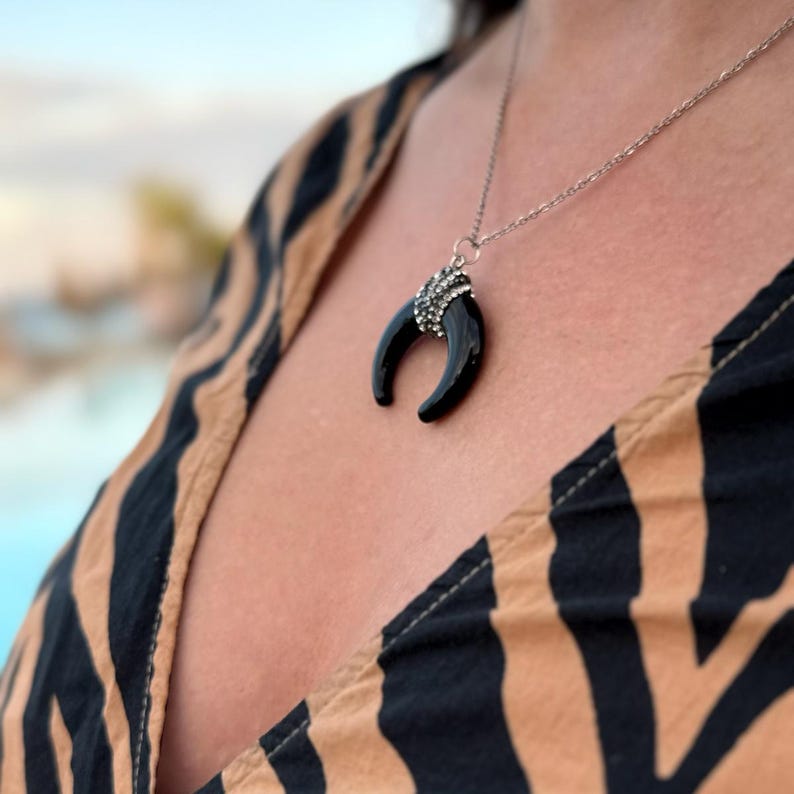 May include: A silver-toned chain necklace featuring a black crescent-shaped pendant. The top of the pendant is embellished with a cluster of small, clear, sparkling stones that catch the light, creating a contrast against the smooth black material.