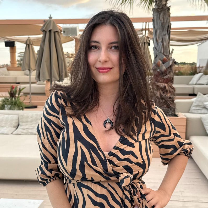 May include: A wrap dress featuring a tan and black zebra print pattern with three-quarter length sleeves and a side tie closure. A silver-toned necklace with a black crescent moon pendant rests against the neckline of the garment.