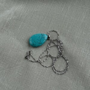 May include: A turquoise teardrop pendant necklace with a silver chain. The pendant is a vibrant blue-green color with dark veining. The chain is delicate and has a textured appearance. The necklace is displayed on a gray fabric surface.
