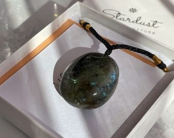 Puede incluir: Un collar de cordón negro con un cierre dorado sostiene un colgante de piedra de labradorita lisa de color gris oscuro. El colgante tiene destellos iridiscentes azules y verdes. El collar se muestra en una caja blanca con el texto "Stardust Nature" impreso en la tapa.