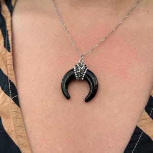 May include: A silver-toned chain necklace featuring a crescent-shaped black pendant. The top of the crescent is adorned with a cluster of small, sparkling clear crystals, creating a contrast between the smooth black surface and the metallic accents.