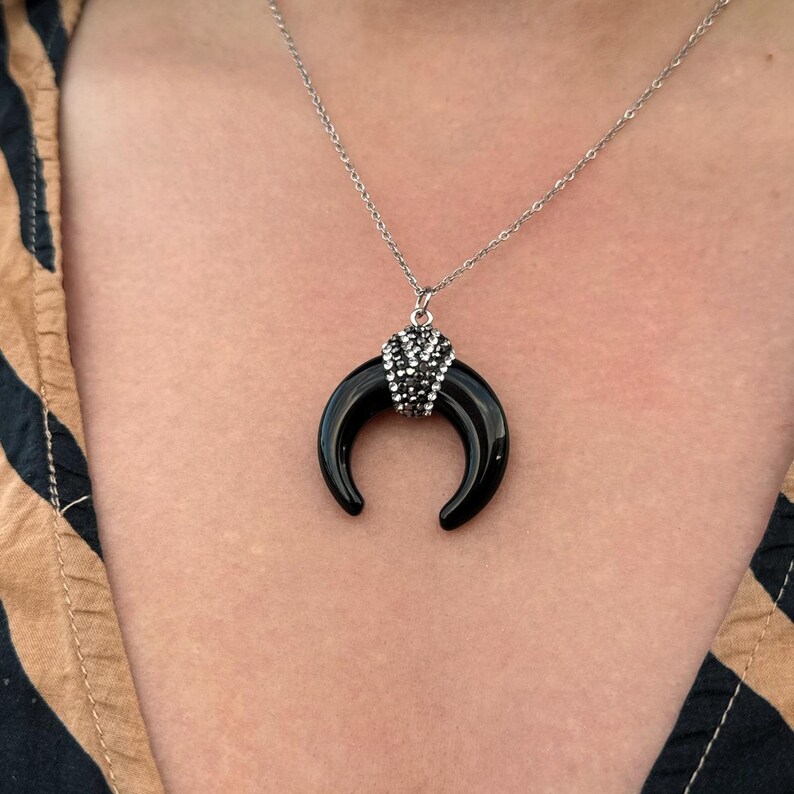 May include: A silver-toned chain necklace featuring a crescent-shaped black pendant. The top of the crescent is adorned with a cluster of small, sparkling clear crystals, creating a contrast between the smooth black surface and the metallic accents.