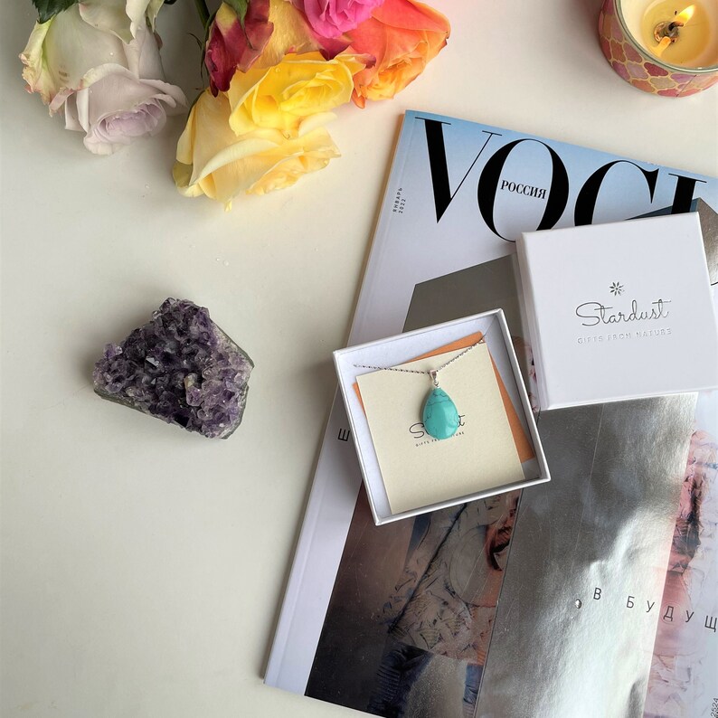 May include: A turquoise teardrop pendant necklace displayed in a white box with the brand name "Stardust". The necklace is on a silver chain. The image also includes a purple amethyst cluster, a magazine, and colorful roses.