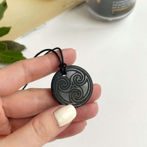 Puede incluir: Collar con colgante negro redondo con un dise&ntilde;o de nudo celta. El colgante est&aacute; hecho de piedra negra y tiene un acabado mate.