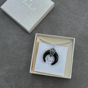 May include: A silver-tone chain necklace featuring a black crescent moon pendant with a sparkling crystal-encrusted cap. The jewelry is presented in a white gift box with a card printed with the text Stardust Gifts From Nature.