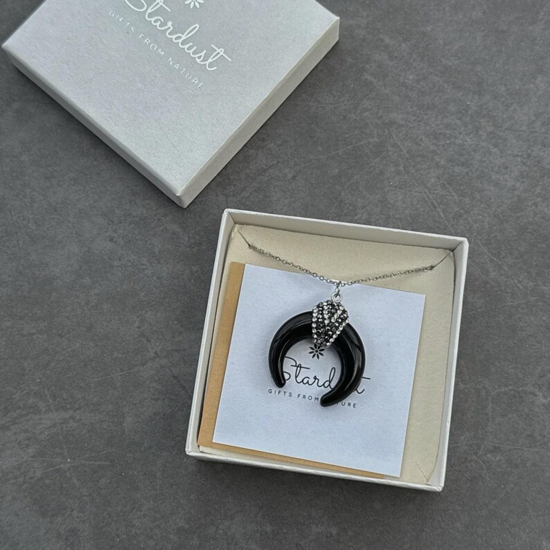 May include: A silver-tone chain necklace featuring a black crescent moon pendant with a sparkling crystal-encrusted cap. The jewelry is presented in a white gift box with a card printed with the text Stardust Gifts From Nature.