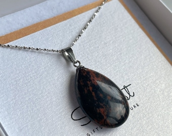 May include: A silver chain necklace with a teardrop-shaped pendant made of red and black jasper. The pendant is polished and has a smooth surface. The necklace is displayed in a white box with a brown paper insert.