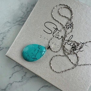 May include: A silver necklace with a large, teardrop-shaped turquoise pendant. The pendant has a marbled appearance with dark blue veins. The necklace is resting on a light gray box with the word "Stardust" printed on it.