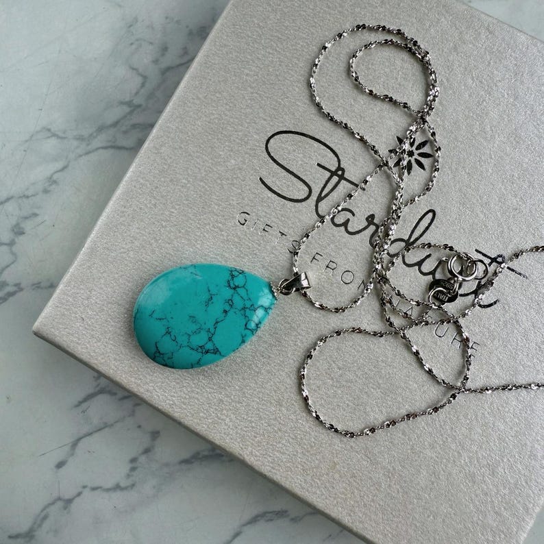 May include: A silver necklace with a large, teardrop-shaped turquoise pendant. The pendant has a marbled appearance with dark blue veins. The necklace is resting on a light gray box with the word "Stardust" printed on it.