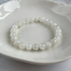 Puede incluir: Un brazalete de cuentas blancas compuesto por piedras redondas y pulidas. El brazalete descansa sobre un plato blanco con borde festoneado. Las cuentas tienen un brillo sutil e iridiscente, lo que sugiere un material natural.