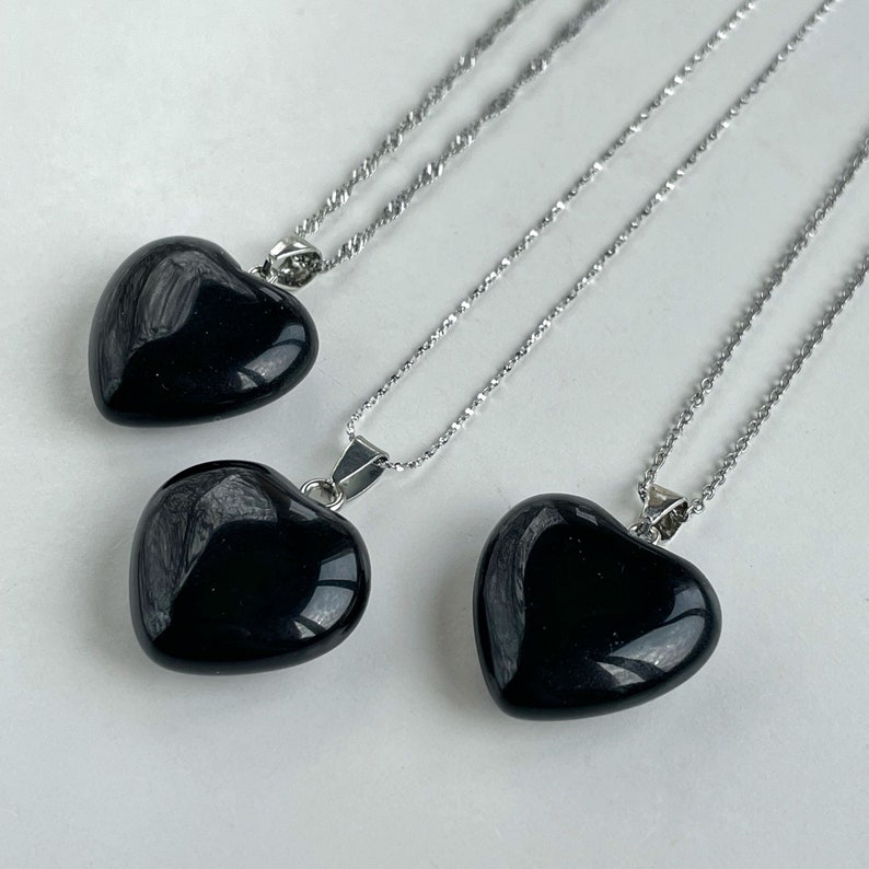 May include: Three necklaces featuring polished, smooth, black heart-shaped pendants suspended from thin, silver-toned metal chains. Each pendant is securely attached to the chain with a small, metallic bail, showcasing a glossy, reflective surface.