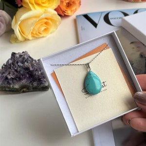 May include: A silver necklace with a teardrop-shaped turquoise pendant is displayed in a white box. The box contains a cream-colored card with the text "Secret Gifts of Nature". The background includes flowers and a purple crystal.