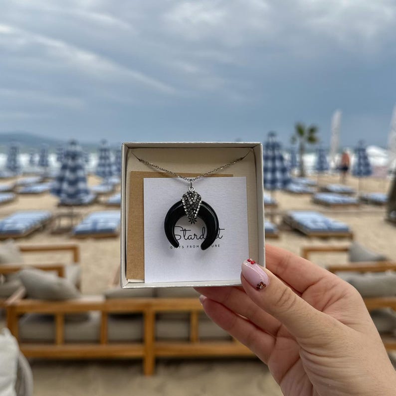 May include: A silver-toned necklace featuring a black crescent moon pendant with a small cluster of clear crystals at the top. The necklace is presented on a white card with the text Stardust Gifts from Nature inside a white box.