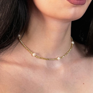 Peut inclure: Un collier ras-du-cou délicat composé d'une chaîne continue de petites perles dorées polies, entrecoupée de trois perles blanches lisses régulièrement espacées. Le collier repose près du cou, présentant un design simple et raffiné.