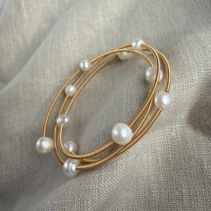 Puede incluir: Una pulsera de alambre dorado con múltiples hebras, adornada con perlas blancas de varios tamaños. La pulsera tiene forma ovalada, colocada sobre una tela de color neutro. Las perlas están espaciadas uniformemente a lo largo del alambre.