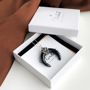 May include: A necklace featuring a black crescent-shaped pendant with a silver-toned cap encrusted with clear crystals, resting inside a white square gift box labeled with the text Stardust Gifts From Nature in a black script font.