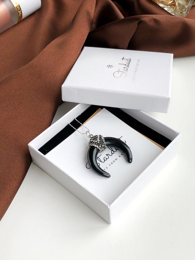 May include: A necklace featuring a black crescent-shaped pendant with a silver-toned cap encrusted with clear crystals, resting inside a white square gift box labeled with the text Stardust Gifts From Nature in a black script font.
