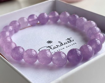 Puede incluir: Un brazalete de amatista púrpura con cuentas redondas en una caja blanca con el texto "Stardust Gifts from Nature".