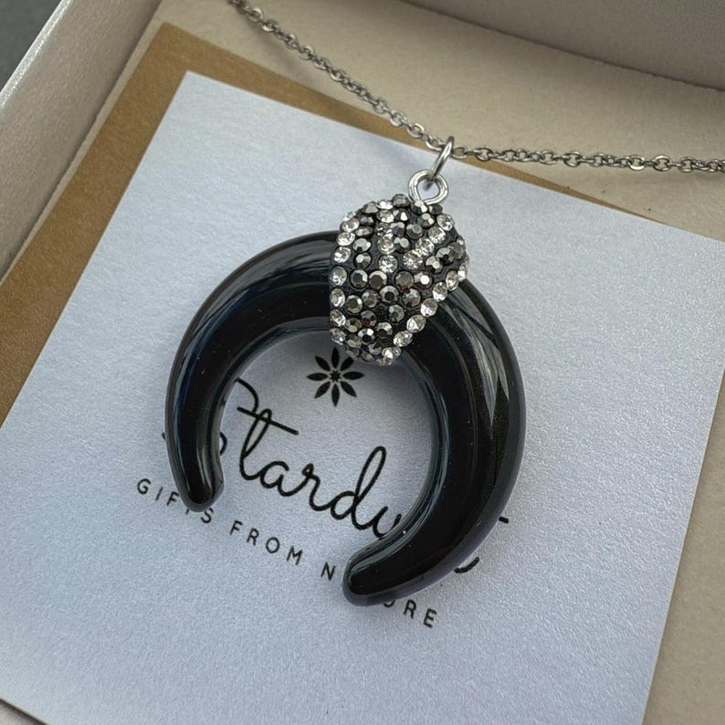 May include: A crescent-shaped black pendant necklace featuring a silver-toned bail encrusted with small clear crystals. The pendant hangs from a thin silver-toned chain and rests on a white card printed with the text Stardust Gifts From Nature.