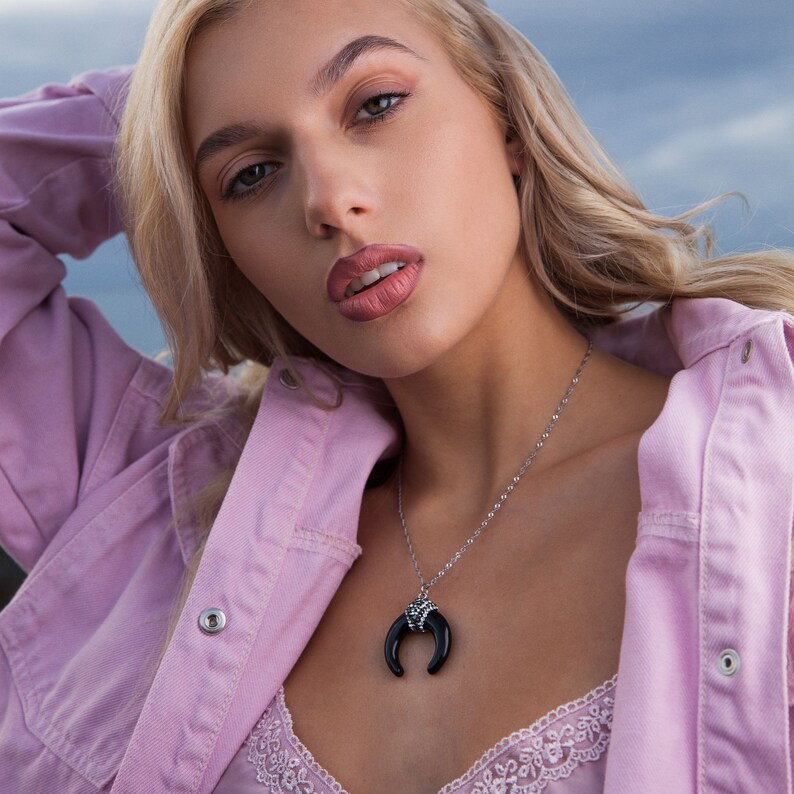 May include: A silver-toned chain necklace featuring a black, crescent-shaped horn pendant with a decorative, textured silver-toned cap at the top. The pendant hangs from a delicate link chain, resting against a light pink lace-trimmed garment.