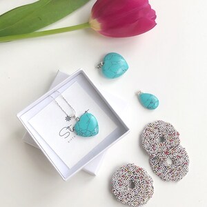 May include: A turquoise heart-shaped pendant necklace in a white gift box, with two loose turquoise pendants and two chocolate-covered cookies with colorful sprinkles. A pink tulip and a sprig of green leaves are also in the image.