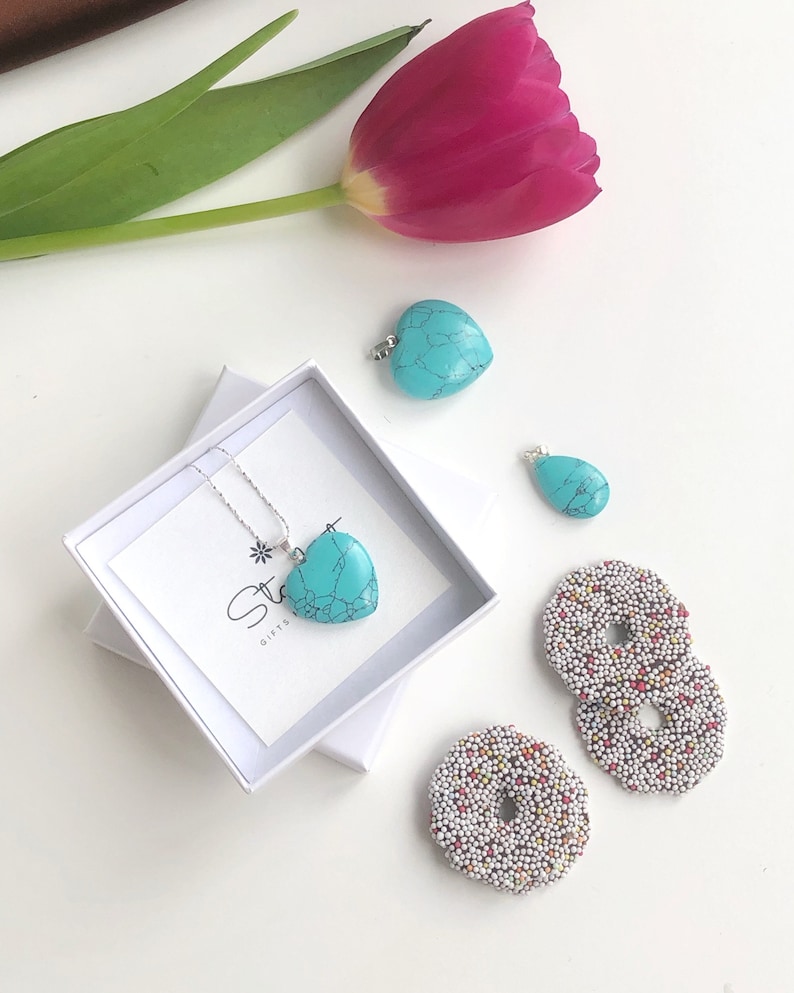 May include: A turquoise heart-shaped pendant necklace in a white gift box, with two loose turquoise pendants and two chocolate-covered cookies with colorful sprinkles. A pink tulip and a sprig of green leaves are also in the image.
