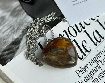 May include: A silver chain necklace with a heart-shaped pendant made of tiger's eye stone. The stone has a brown, gold, and black pattern. The necklace is on a white surface with text that reads "Stardust Gifts from Nature".