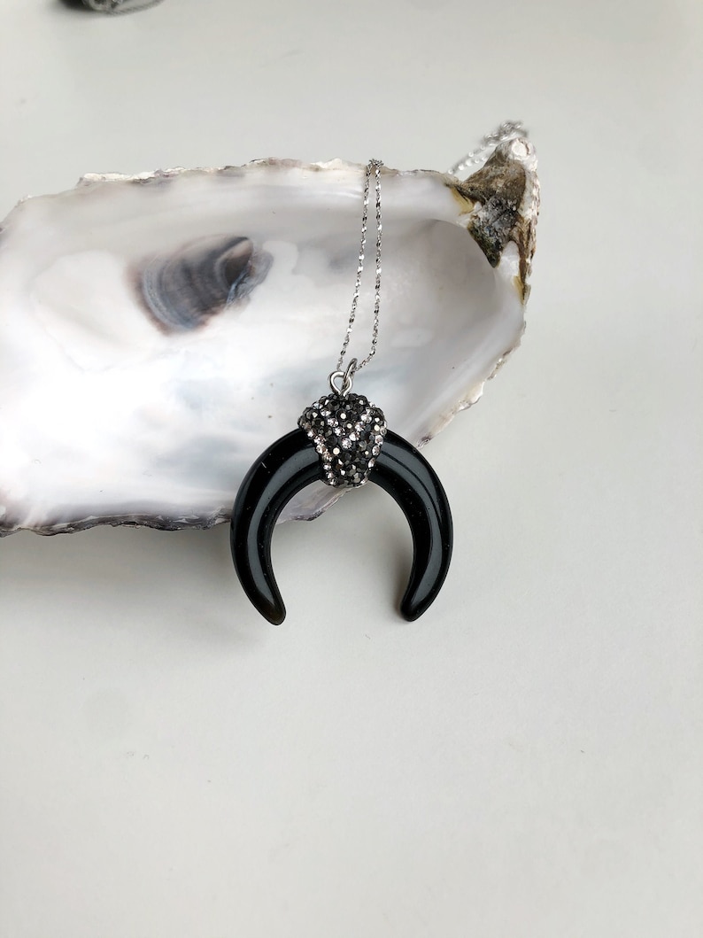 May include: A necklace featuring a smooth, black, crescent-shaped pendant suspended from a silver-toned chain. The top of the crescent is adorned with a cluster of small, sparkling black crystals, creating a contrast between the polished surface and the textured, glittering embellishment.