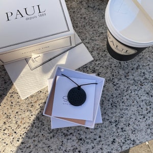 Puede incluir: Un collar negro con un colgante redondo negro en una caja blanca. La caja est&aacute; etiquetada con "Gifts from the Heart".