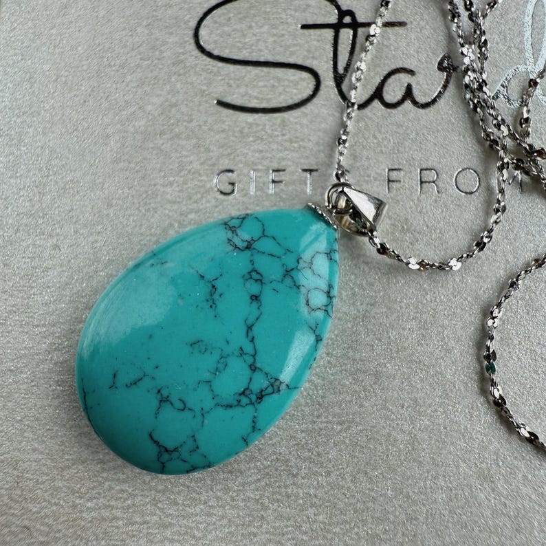May include: A turquoise teardrop pendant necklace with a silver chain. The pendant has dark veining and a polished surface. The necklace is displayed on a gray surface with the text "Star GIFTS FRO" visible.