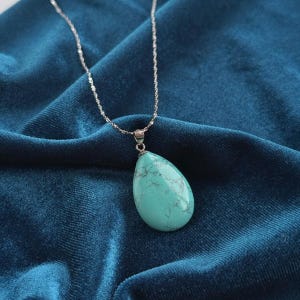 May include: A silver necklace with a teardrop-shaped turquoise pendant. The pendant is a light blue-green color with dark gray veining. The necklace chain is a delicate, twisted design, and the background is a dark blue velvet fabric.