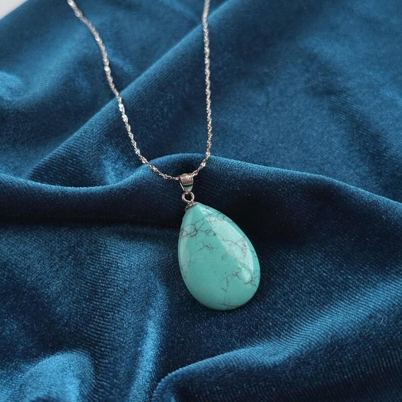 May include: A silver necklace with a teardrop-shaped turquoise pendant. The pendant is a light blue-green color with dark gray veining. The necklace chain is a delicate, twisted design, and the background is a dark blue velvet fabric.